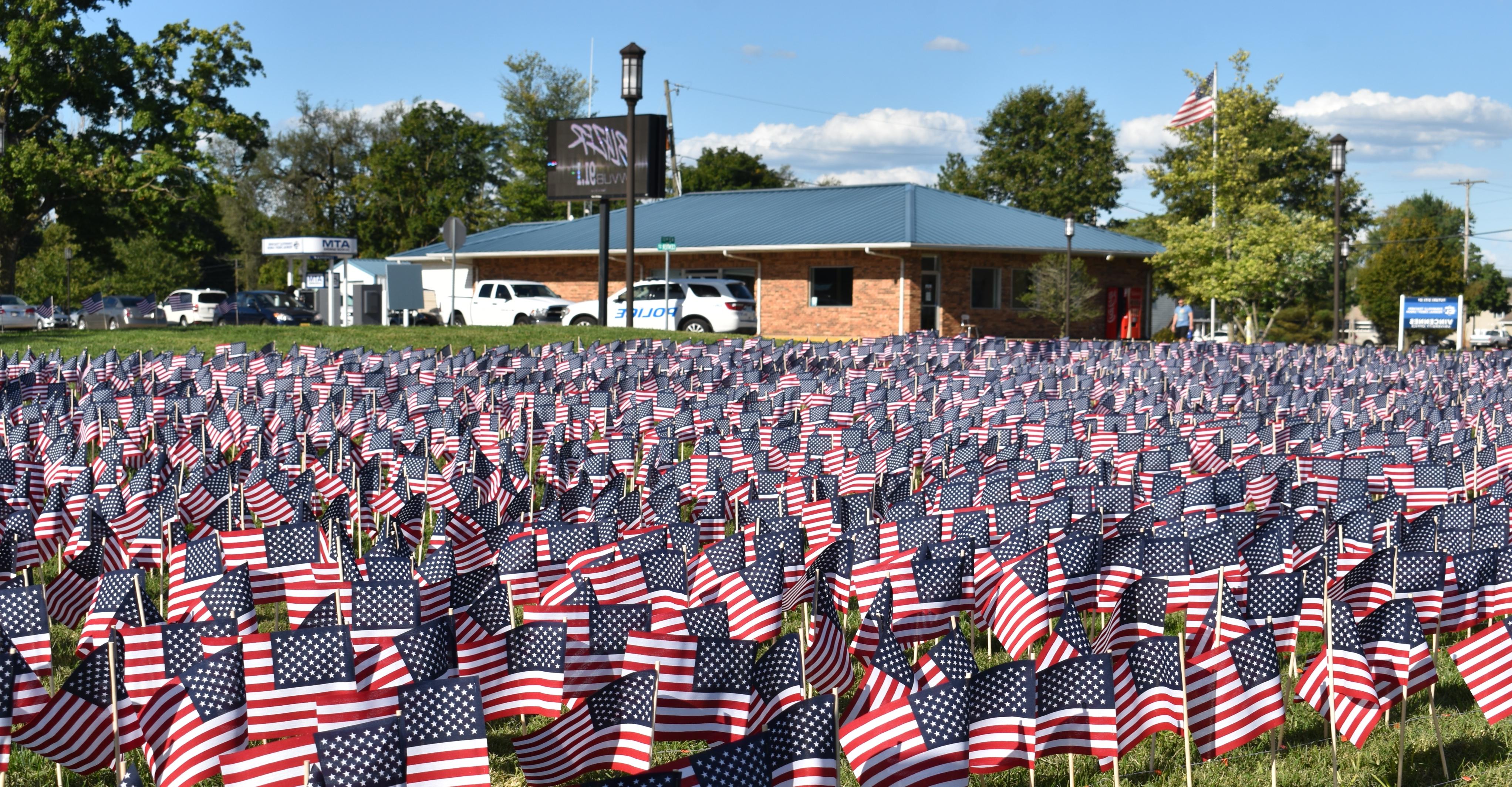 Hundreds of small American flags in a grassy field with a flagpole flying an American flag and the 澳门足球博彩官方网址 Police Department building in the background.